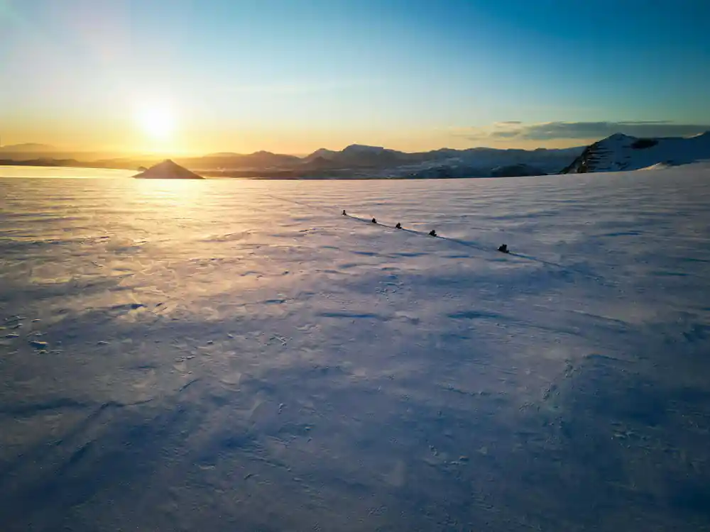 Group of people riding snowmobiles across frozen lake at sunset