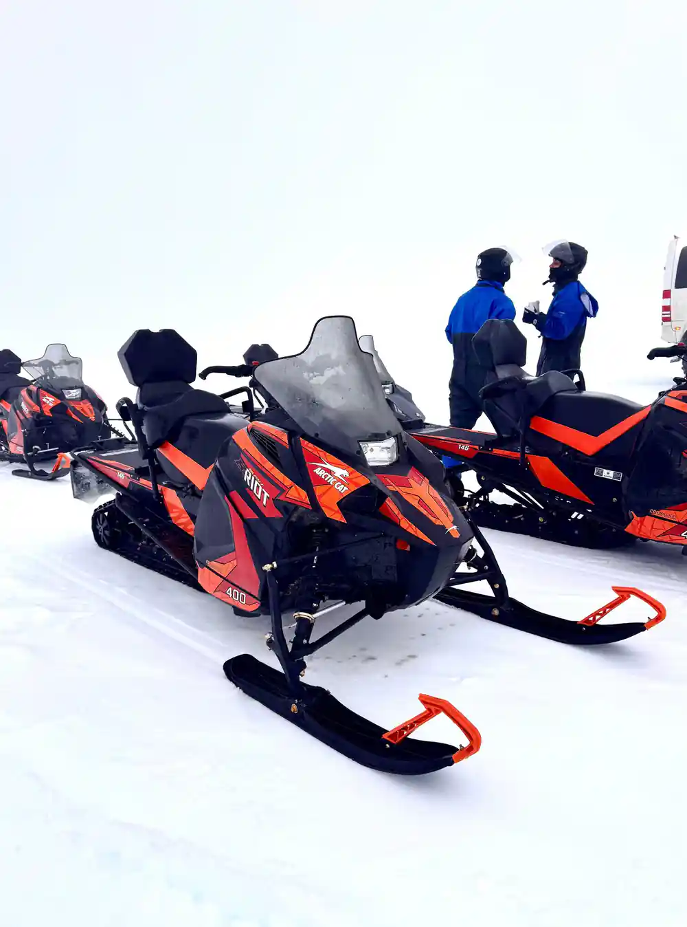 Orange snowmobiles parked on snow with riders standing
