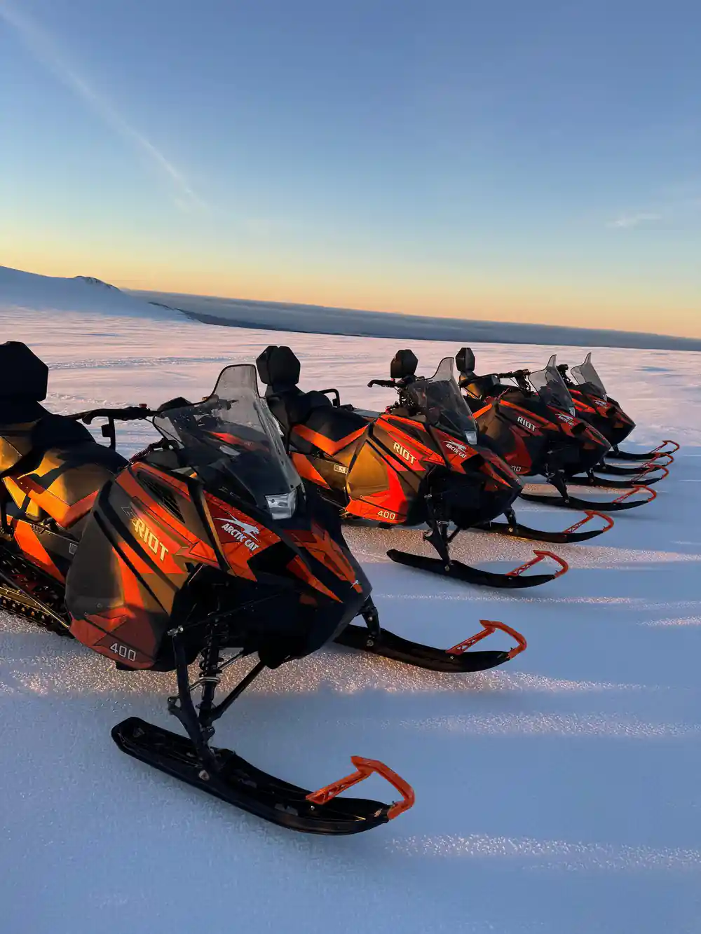 Riders on orange snowmobiles lined up on snow