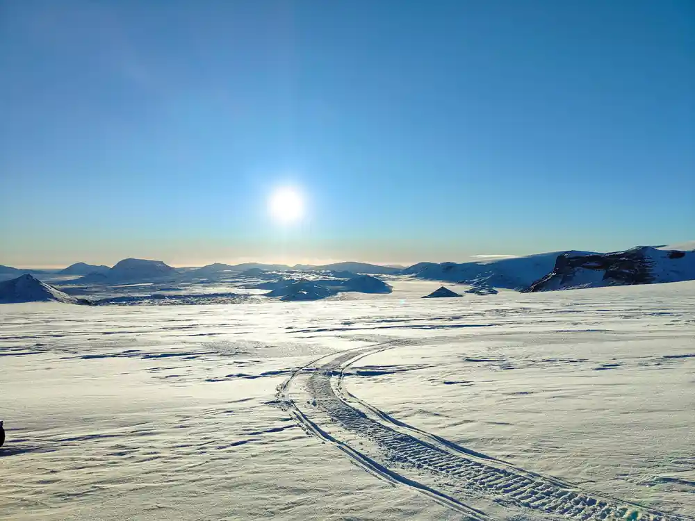 Snowmobile tracks across white landscape with sun and mountains