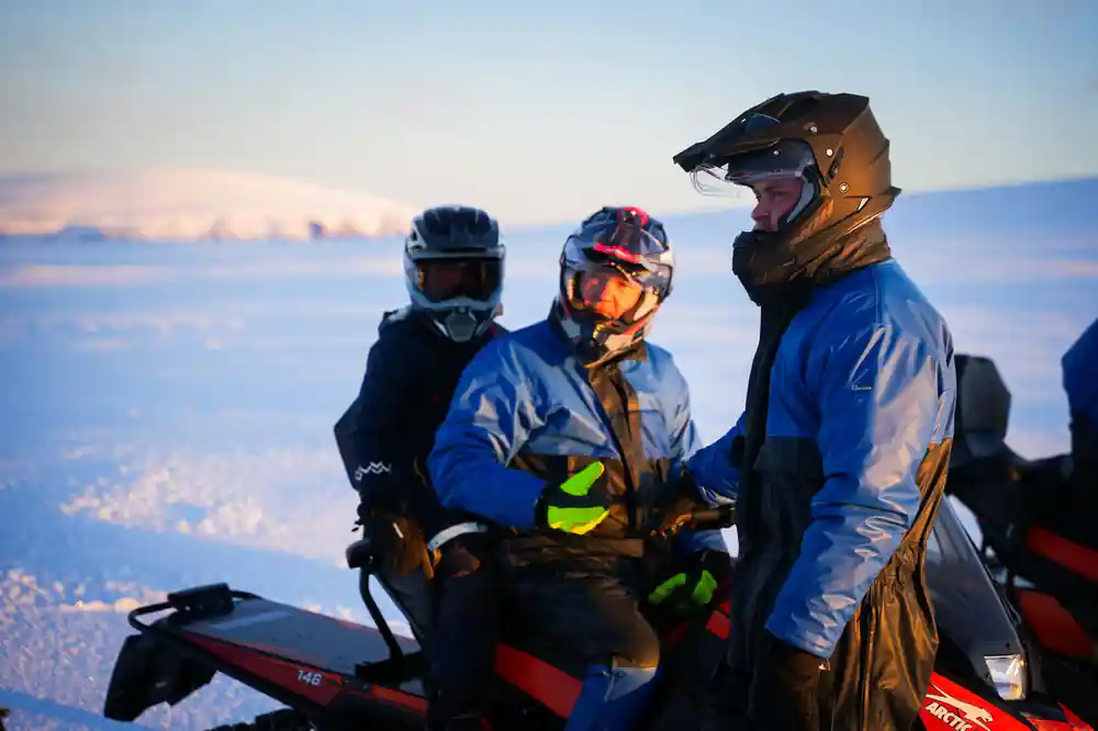 Three snowmobilers in helmets above clouds at sunset