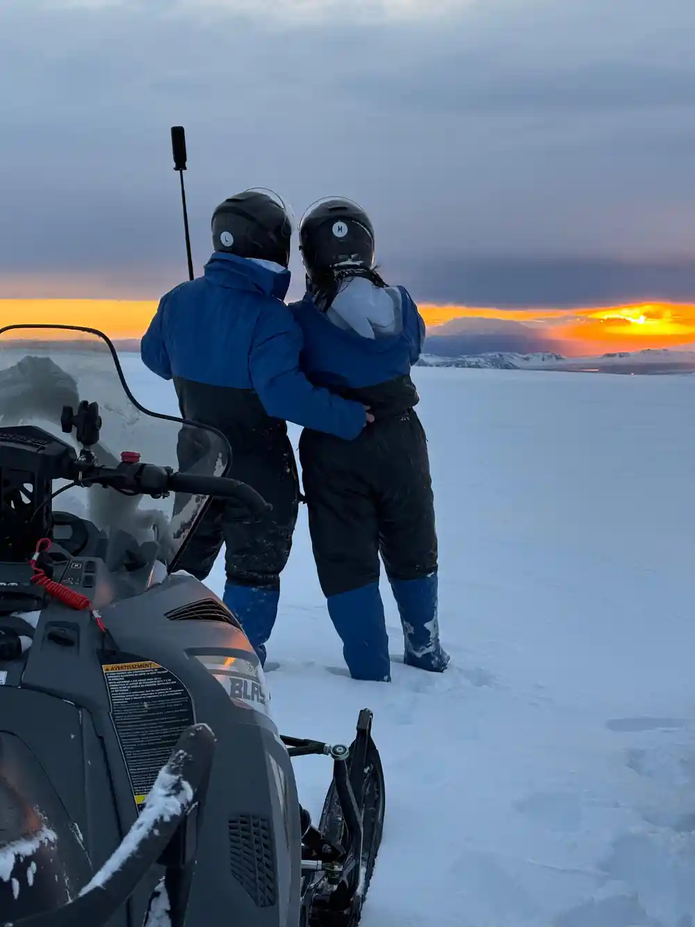 Two people standing on snowmobiles at sunset by frozen water